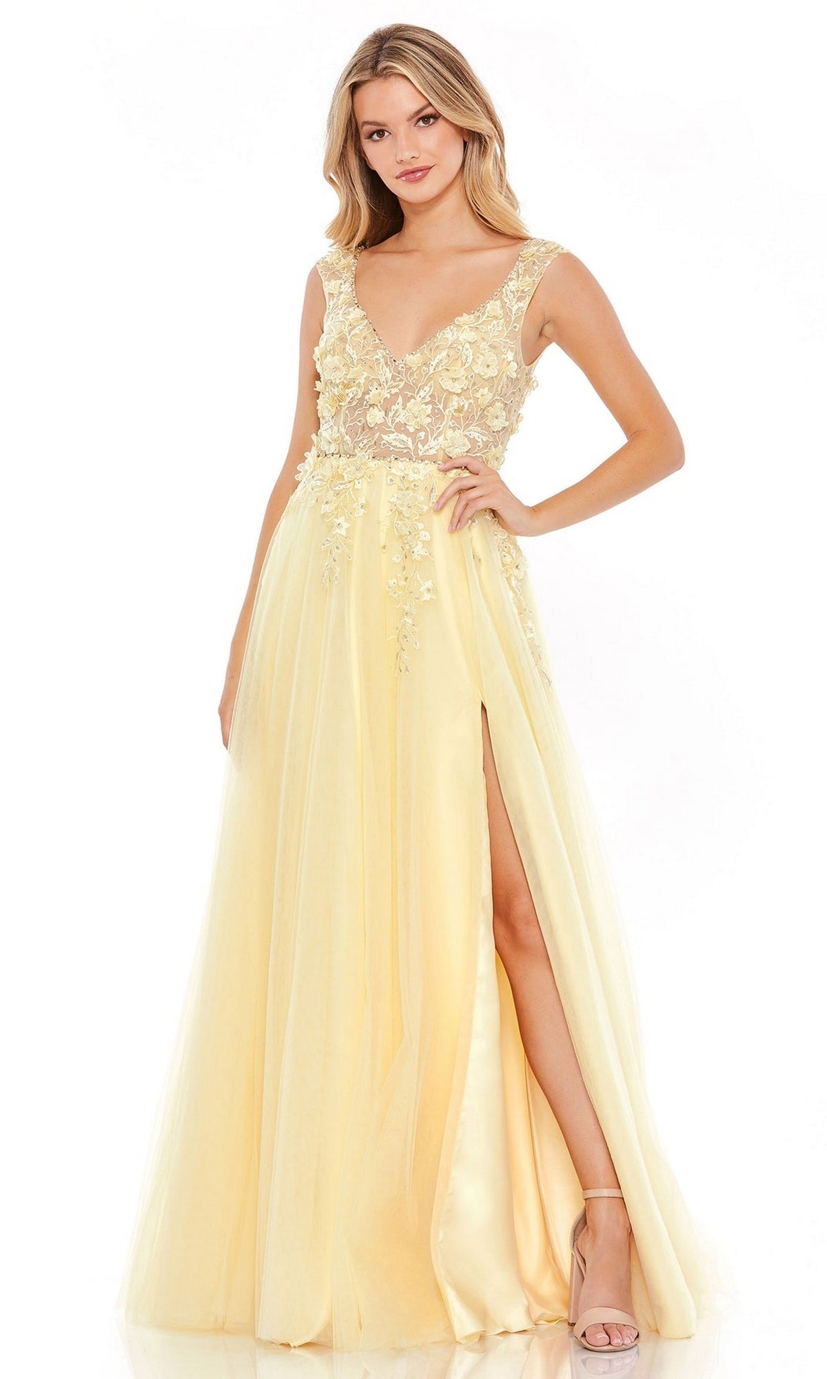 A woman stands confidently with one hand on her hip, wearing the Mac Duggal Long Formal Dress 11201—a yellow A-line gown featuring a sheer bodice, floral embroidery, and a high slit.